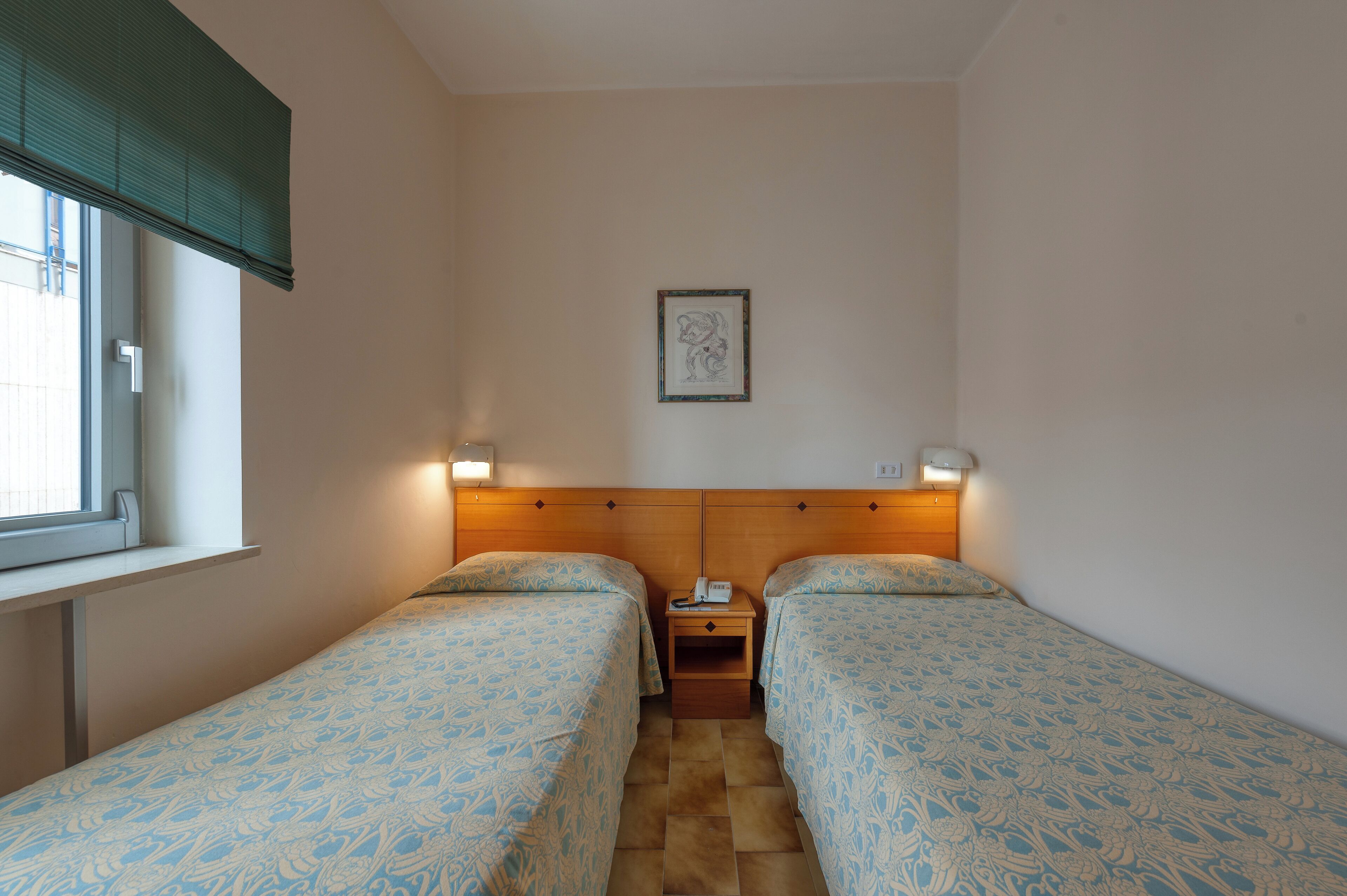 Une petite chambre propre, avec deux lits simples, une table de chevet avec un téléphone et une fenêtre équipée de stores.