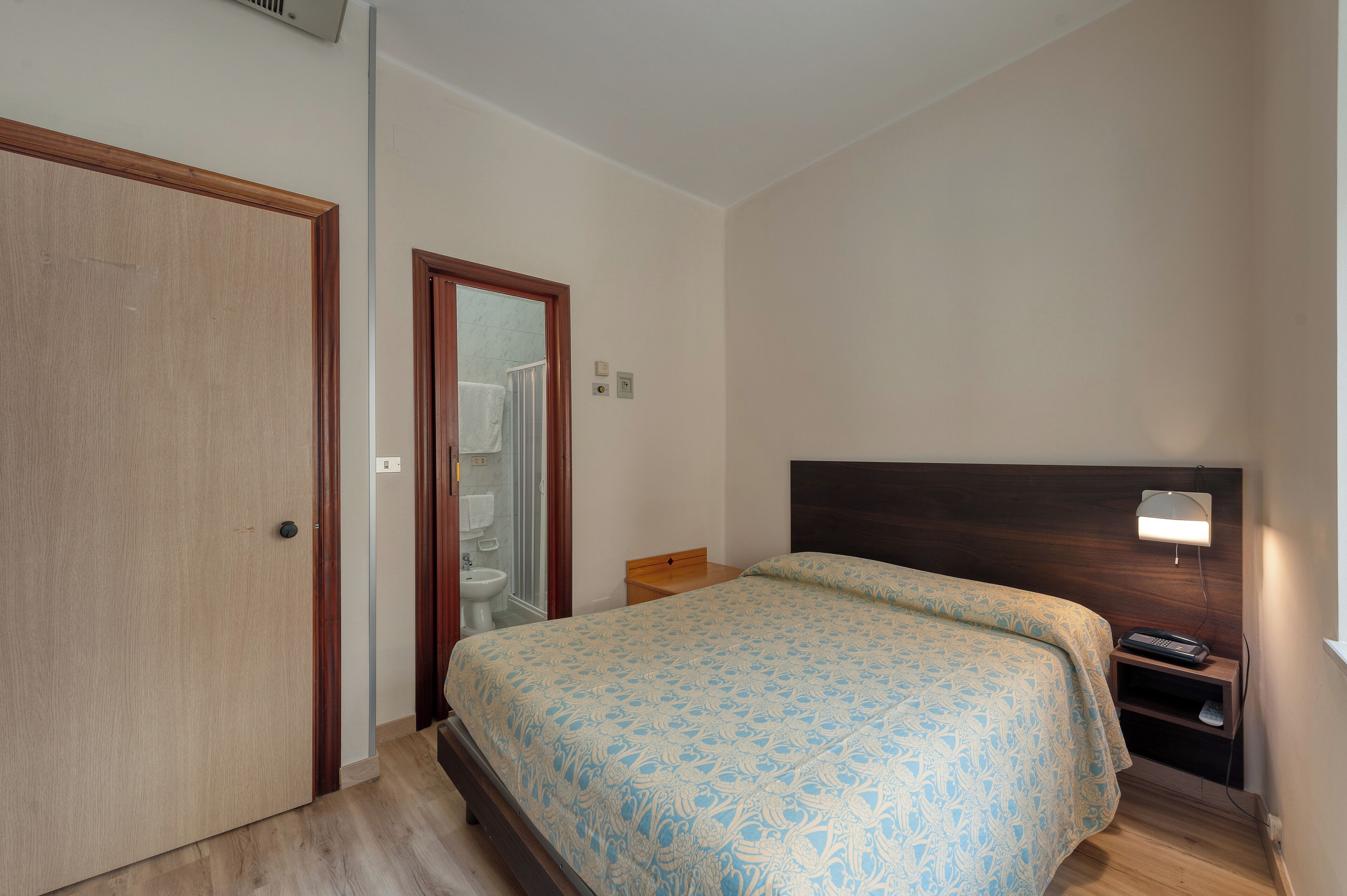 A bedroom with a wooden door, a bedside table, a bed with a patterned bedspread, a nightstand with a lamp, and a small shelf.