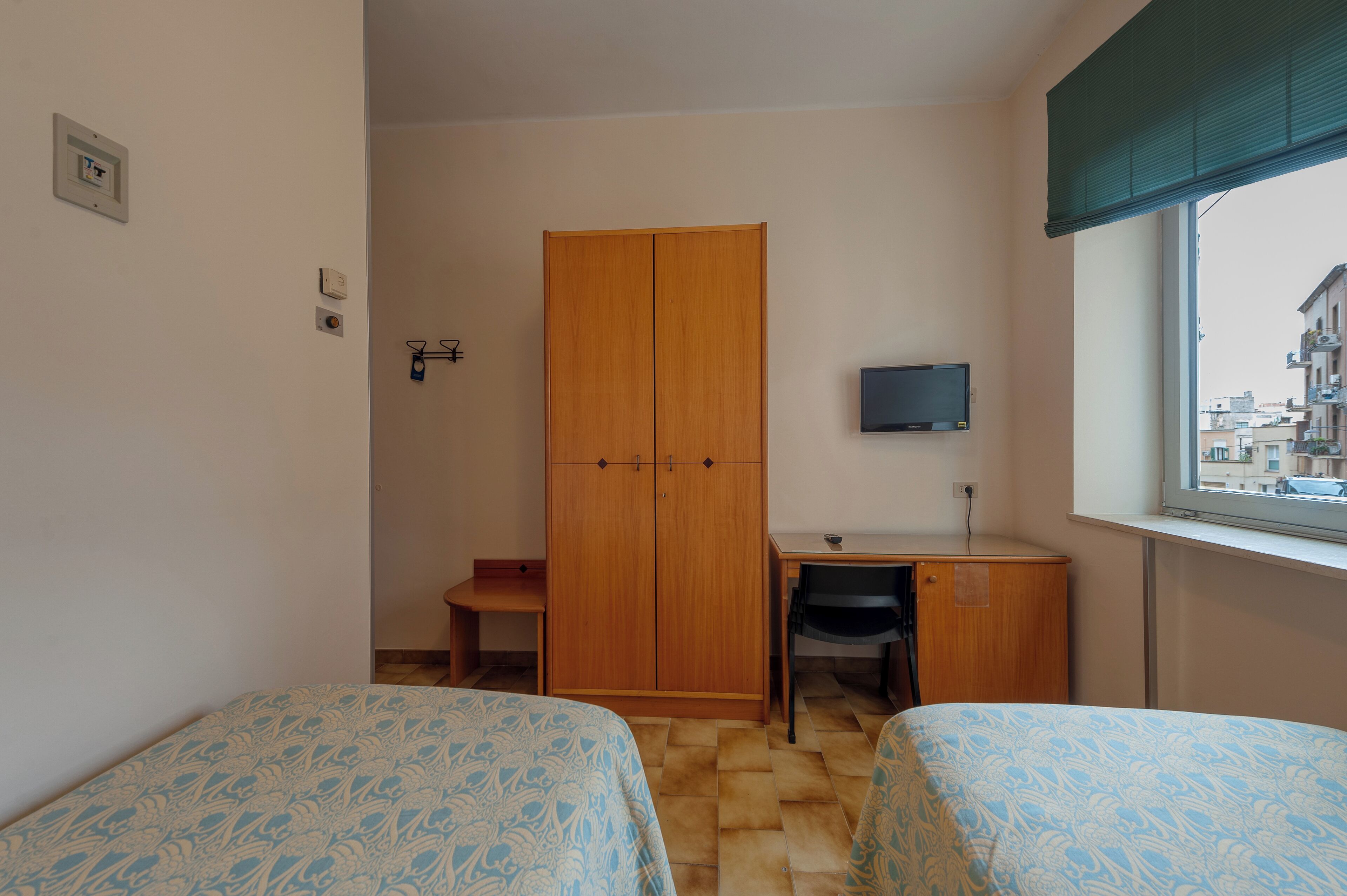 A small room with two beds, a wooden wardrobe, a desk with a chair, a television, and a window with a view of buildings.