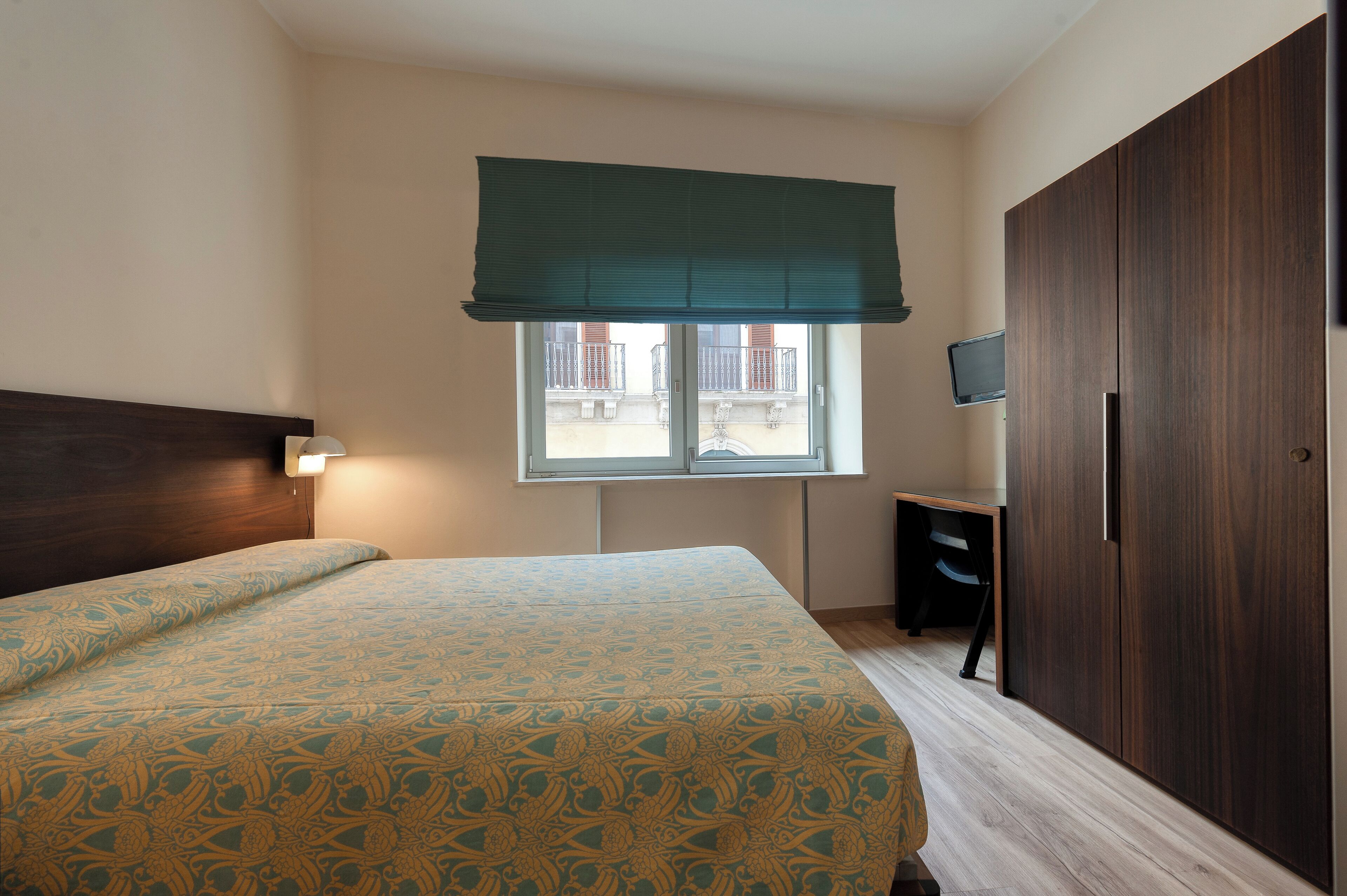 Un dormitorio con cama, escritorio, armario y ventana con vistas a edificios.