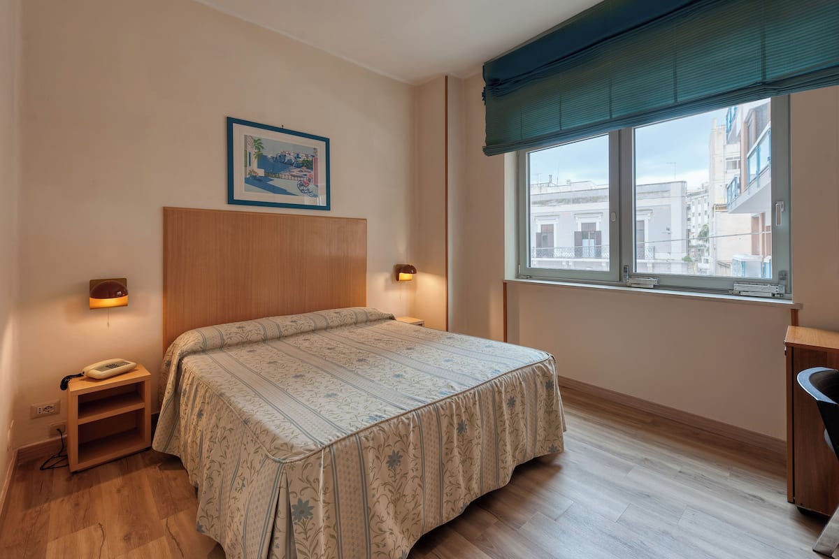 A bedroom with a bed, bedside lamps, a telephone, a small table, and a window with a view of buildings.