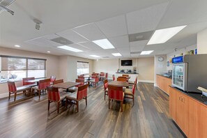 Un comedor amplio con mesas de madera y sillas tapizadas en rojo, una zona de cocina con refrigerador y televisor, y ventanas grandes que ofrecen luz natural.
