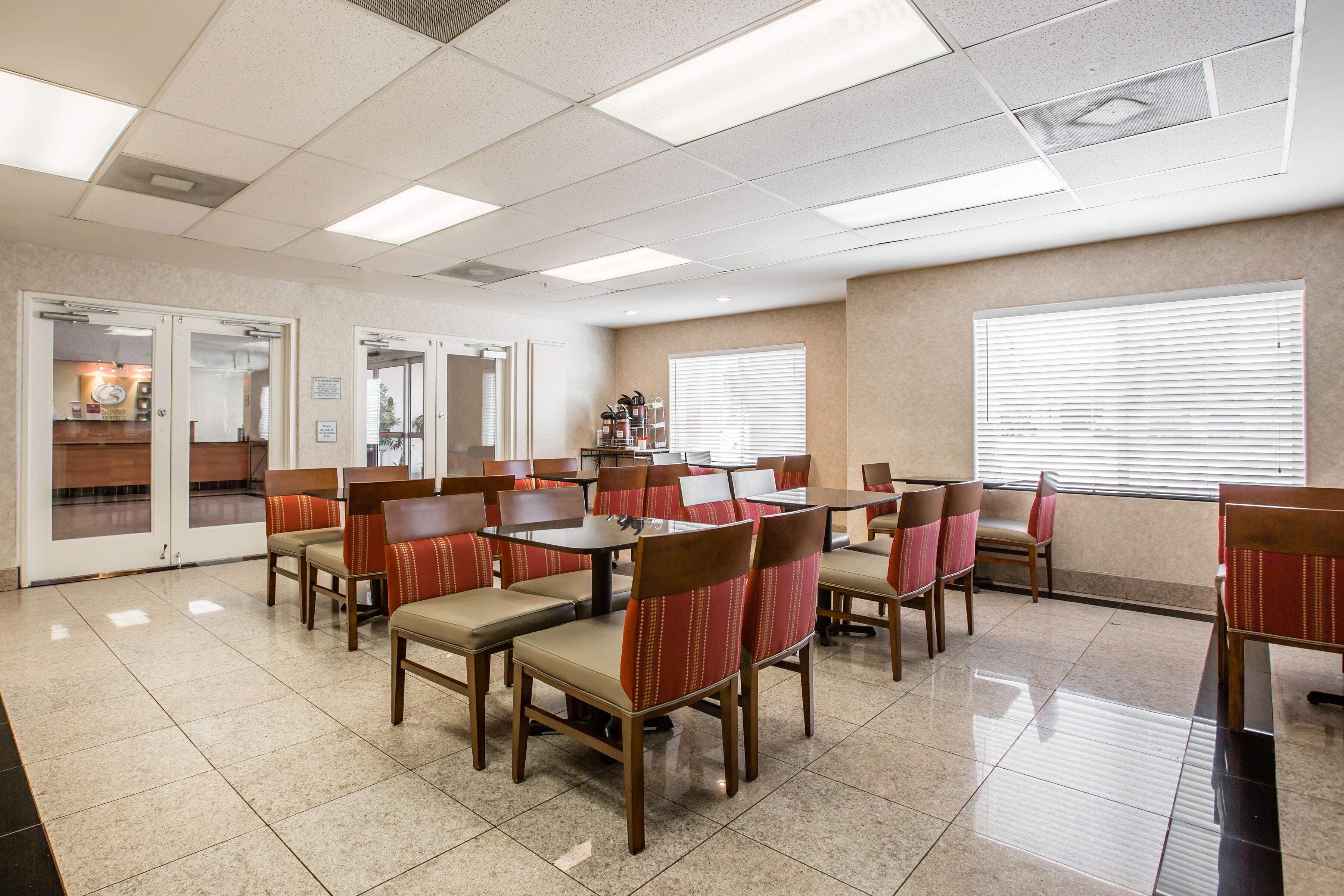 A dining area with tables and chairs, a kitchen in the background, and a window with blinds.