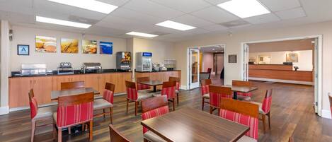 A modern dining area with wooden tables and chairs, a kitchen counter with appliances, and a reception desk.