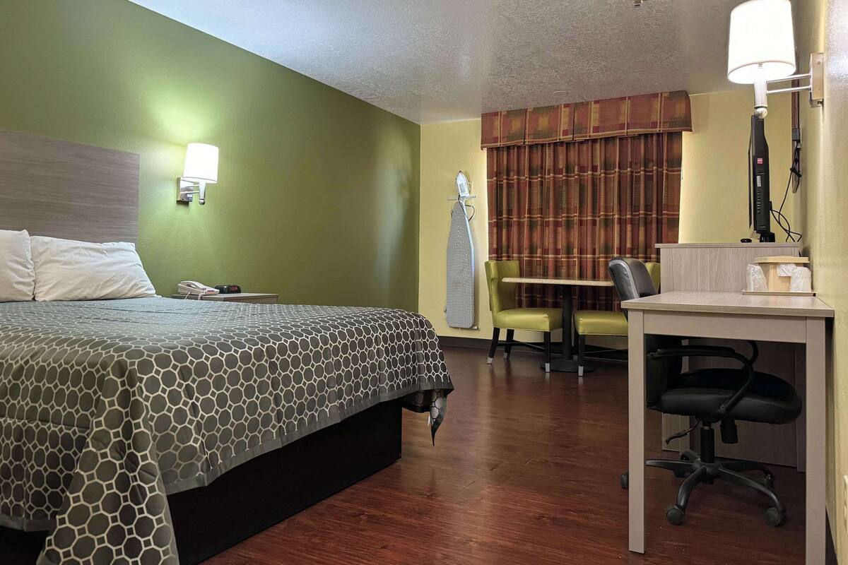 Quarto de hotel com cama, escrivaninha, cadeira, luminária e janela com cortinas.