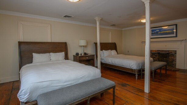 A hotel room with two single beds, a bench, a nightstand with a lamp, and a fireplace. - Bristol Harbor Inn (BRISTOL)