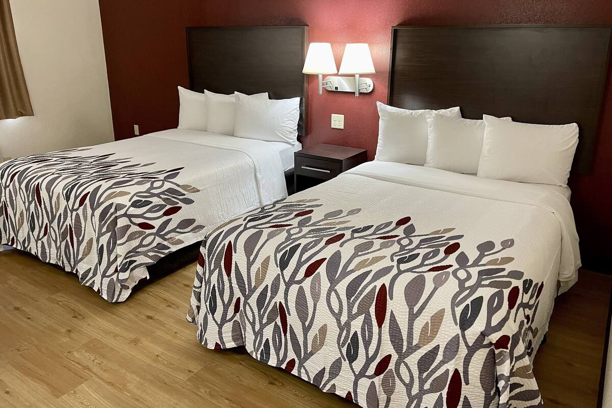 A hotel room with two beds, each with white bedding and a leaf-patterned cover. There are two bedside tables with lamps and a nightstand. The room has a wooden floor and red walls.