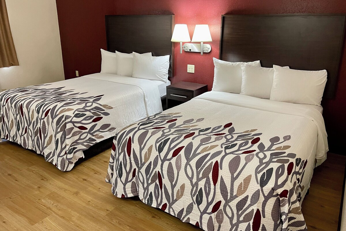 A hotel room with two beds, each with white bedding and a leaf-patterned cover. There are two bedside tables with lamps and a nightstand. The room has a wooden floor and red walls.