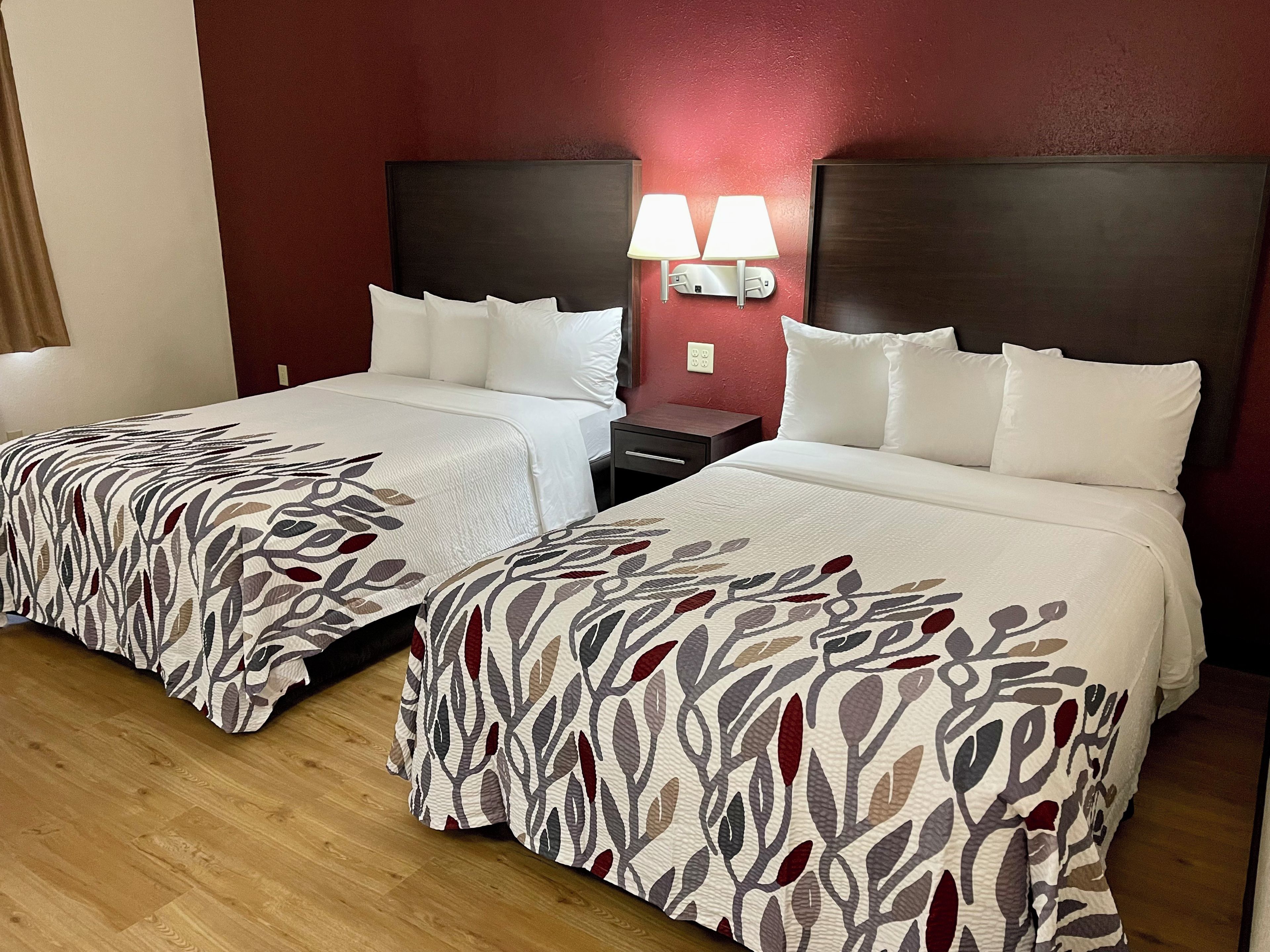 Habitación de hotel con dos camas, cada una con ropa blanca y cubrecama estampado con hojas. Hay dos mesitas de noche con lámparas, y las paredes están pintadas de rojo.