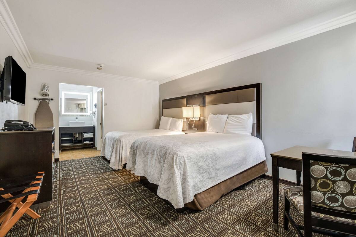 A hotel room with two beds, a desk, a chair, a TV, and a bathroom visible through an open door.