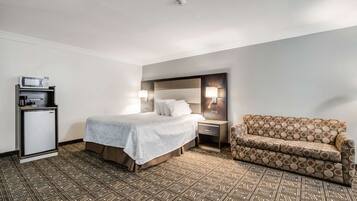 Habitación de hotel con cama, mesita de noche con lámpara, sofá, minibar y alfombra estampada.