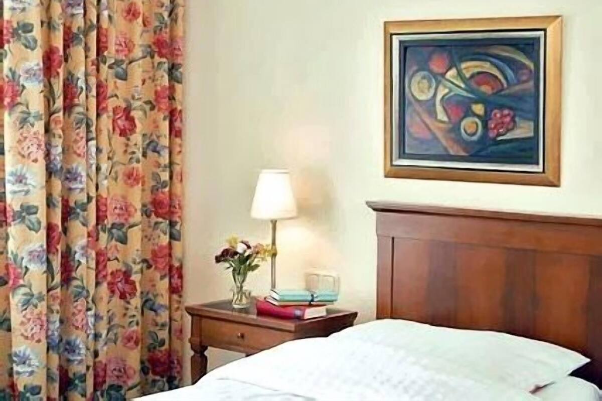 Una habitación de hotel con una cama, una mesita de noche con una lámpara, un jarrón con flores y un cuadro en la pared.