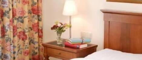 A hotel room with a bed, a nightstand with a lamp, a framed painting, and floral curtains.