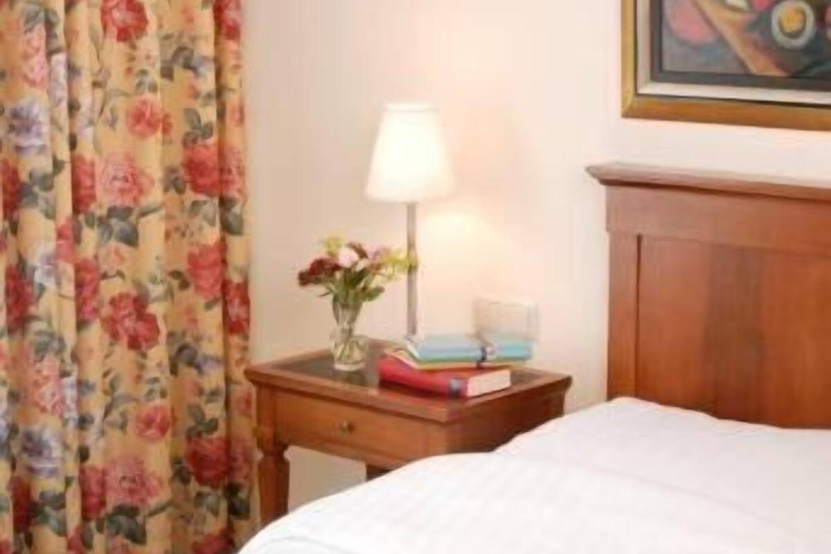 Una habitación de hotel con una cama, una mesita de noche con lámpara, un cuadro enmarcado y cortinas con motivos florales.