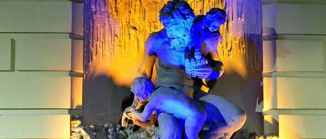 A stone fountain with a statue of a figure holding a child, set in a niche with a decorative arch and illuminated by blue and yellow lights.