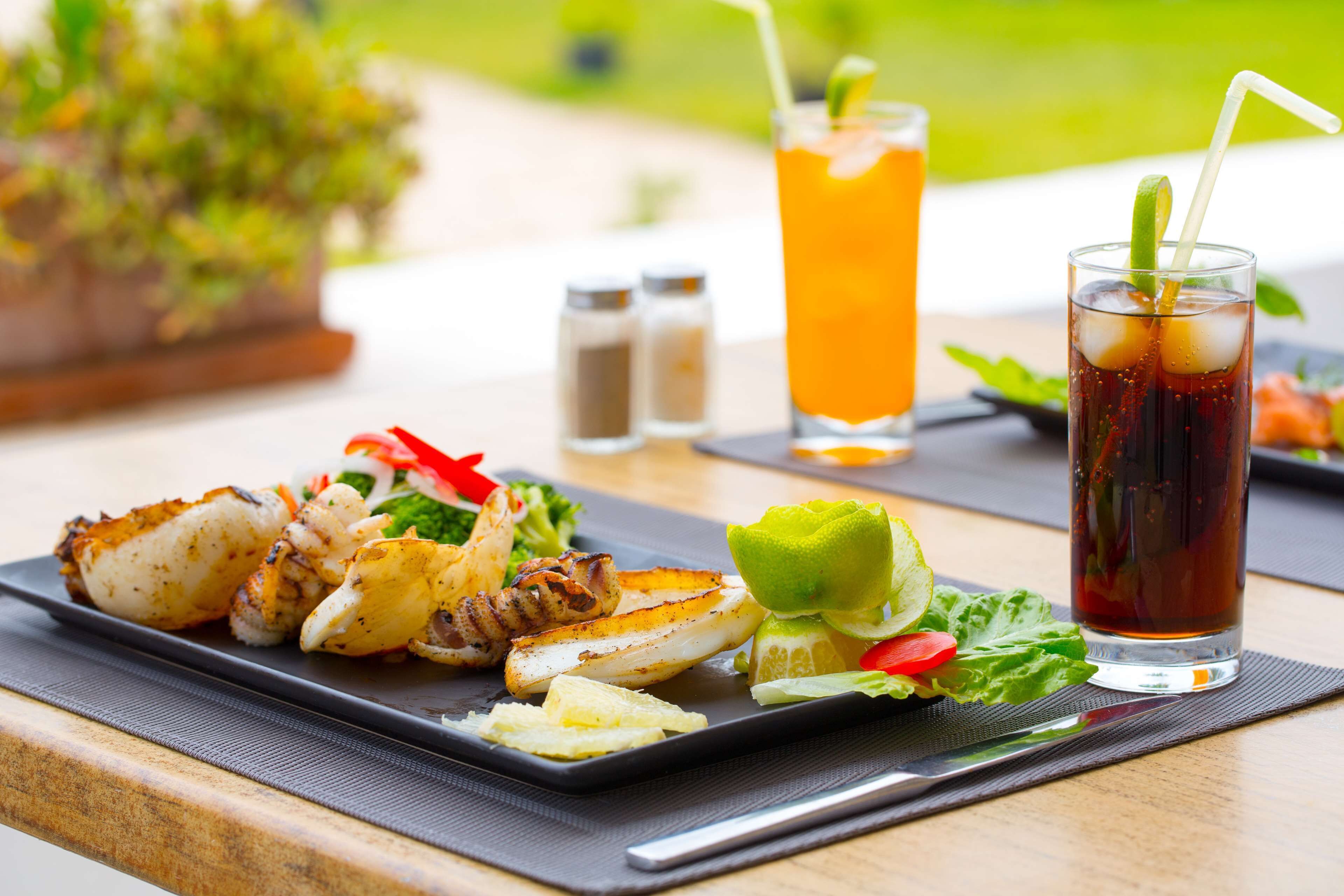 Una comida con mariscos a la parrilla y verduras, un vaso de té helado y una bebida adornada con lima sobre una mesa de madera.