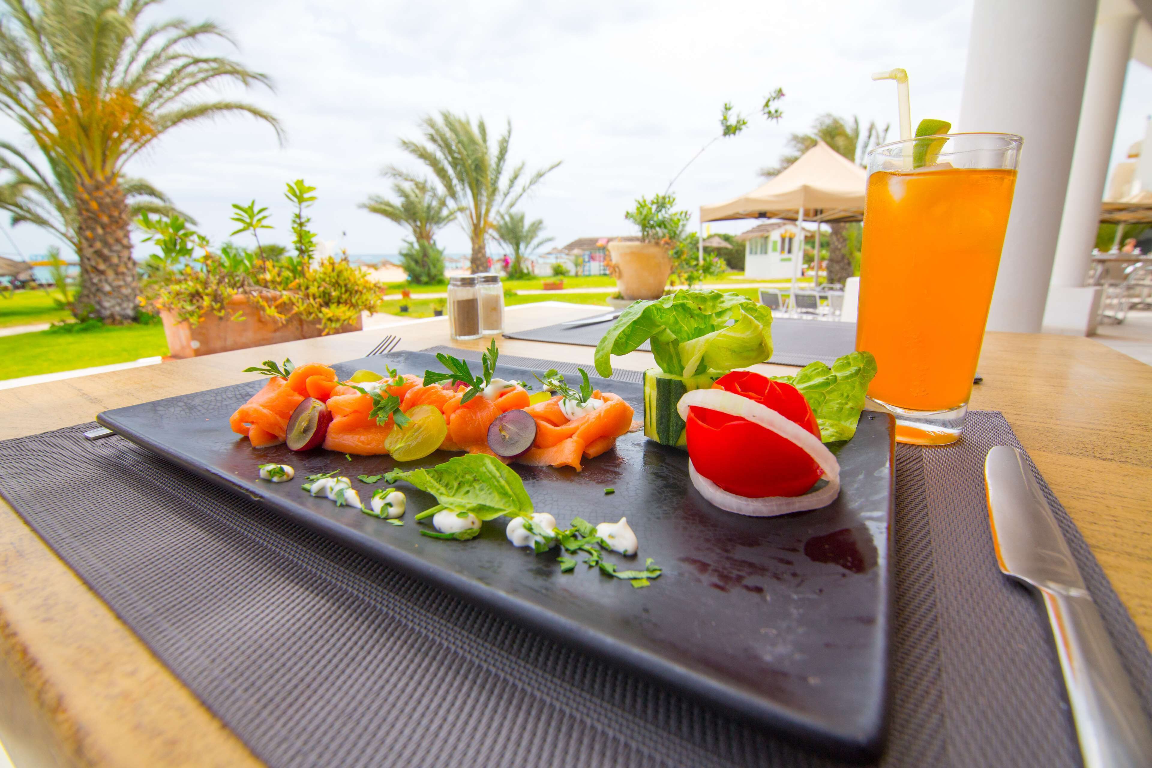 Eine Mahlzeit mit Gemüse und Lachs auf einem schwarzen Teller, ein Glas Orangensaft und ein Messer auf einem Tisch mit Blick auf Palmen und einen Pool.