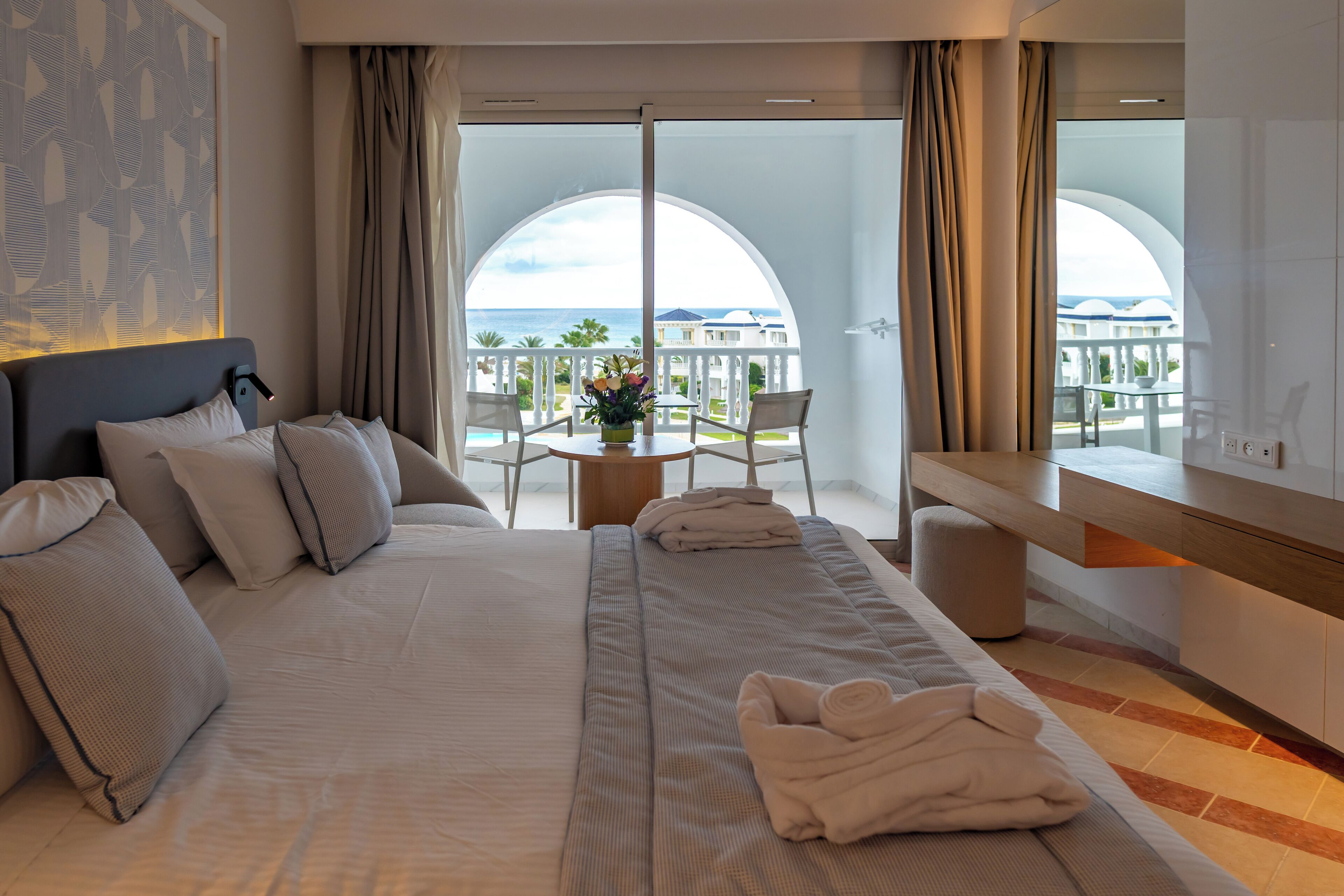 A hotel room with a large bed, a small table with a vase of flowers, and a balcony with a view of the sea.