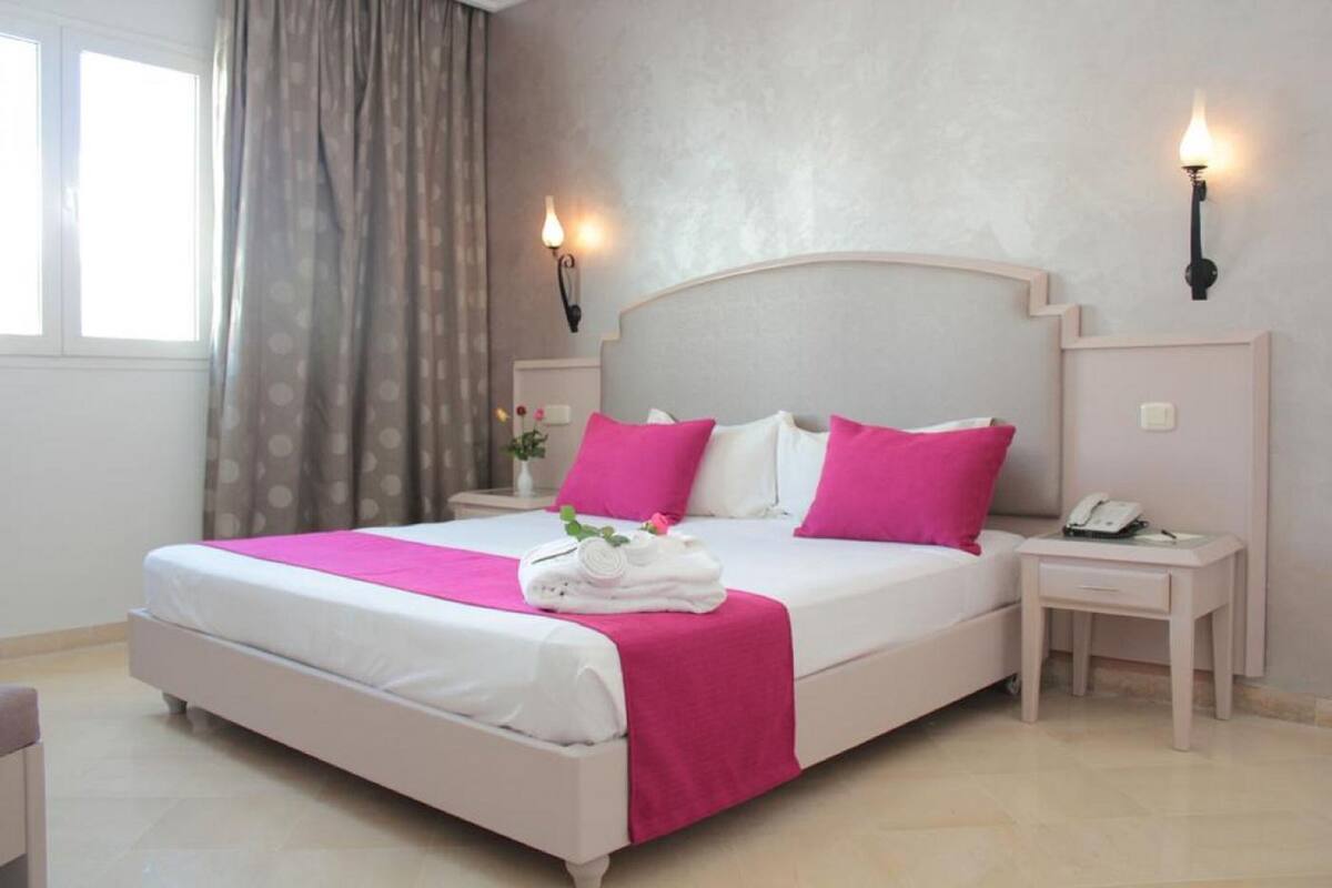 Un lit bien fait, aux couleurs rose et blanc, une table de chevet avec un téléphone et une fenêtre avec des rideaux.