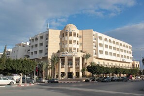 A multi-story hotel with a central atrium, balconies on each floor, and a parking lot in front. - El Kantaoui Center and Spa (Port El Kantaoui)