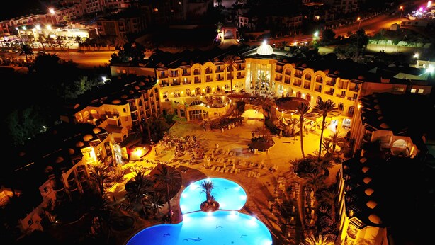 A large, illuminated swimming pool with a central fountain, surrounded by sun loungers and palm trees, set within a resort complex with multiple buildings and a well-lit street in the background. - Eden Star Resort (Zarzis)