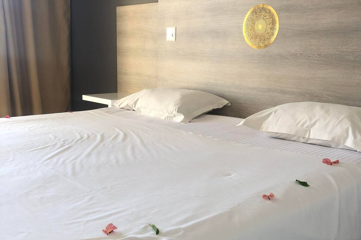 A neatly made bed with white linens and a few decorative flowers, a wooden headboard, and a wall-mounted light fixture.