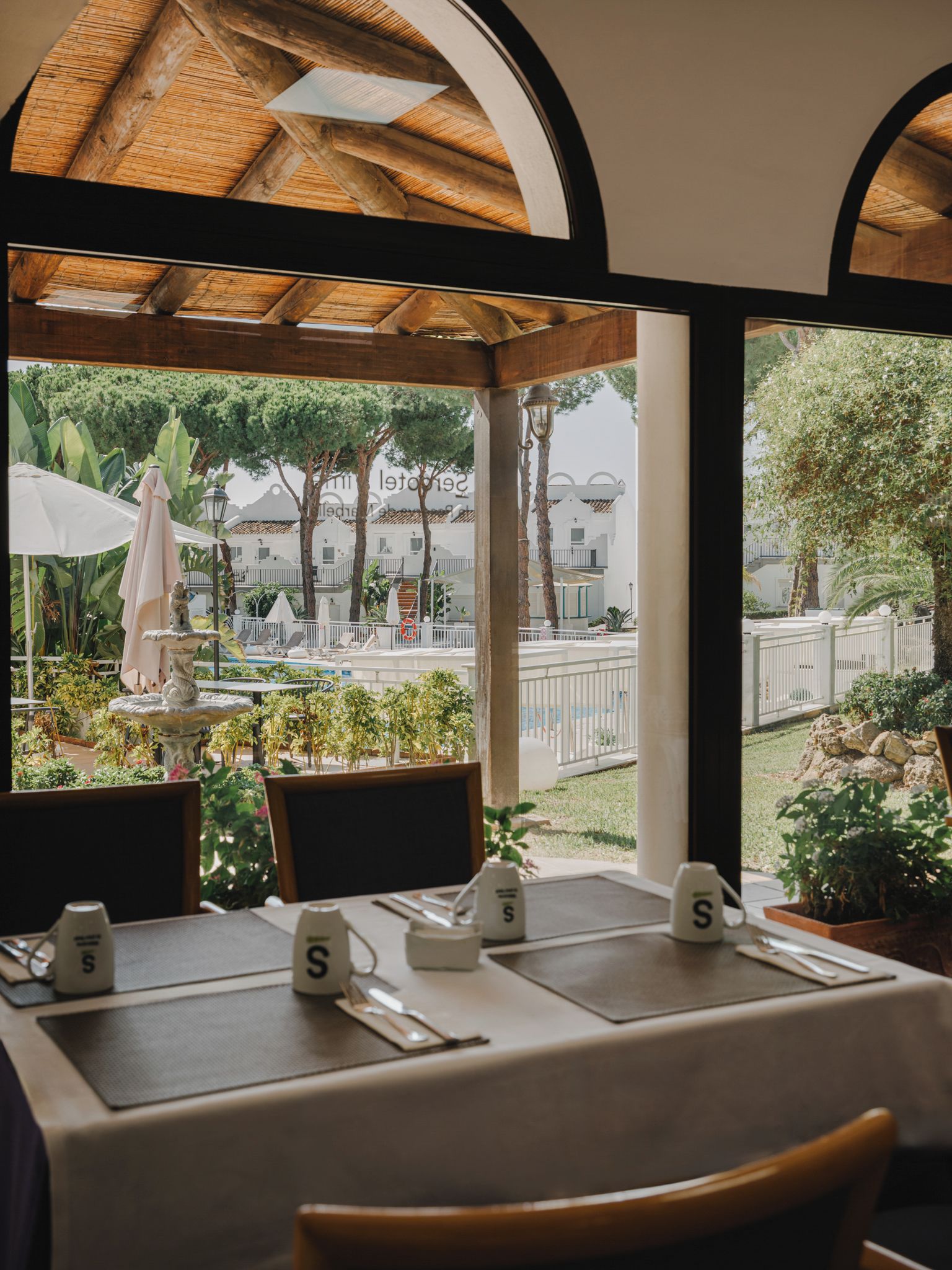 An outdoor dining area with tables set for two, a fountain, and a view of a pool and buildings.