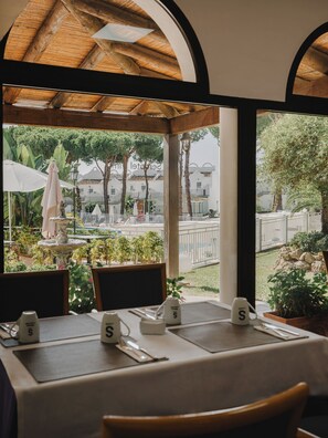Un área de comedor al aire libre con mesas dispuestas para dos, una fuente y vistas a una piscina y edificios.