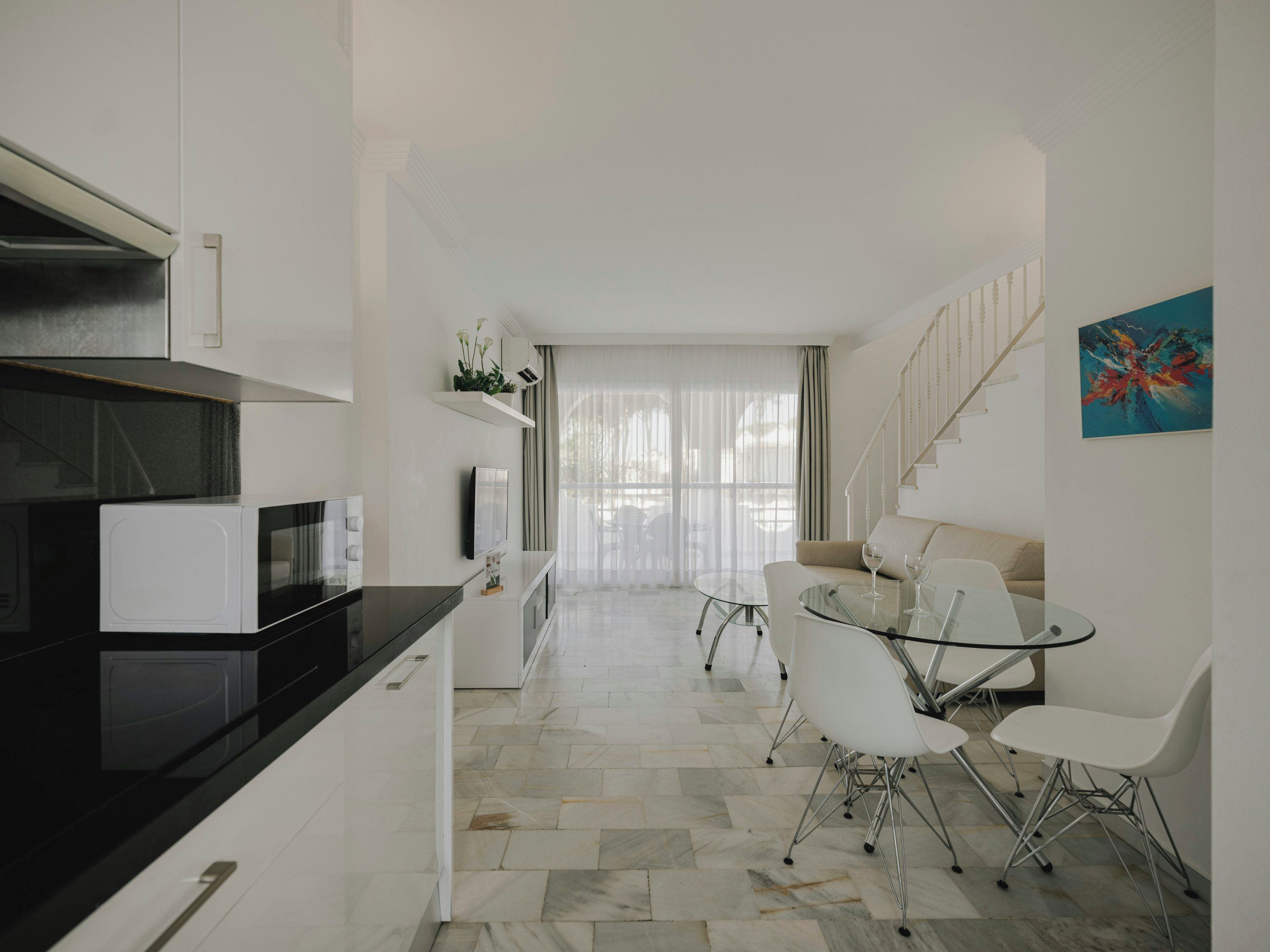 Una cucina moderna con un piano di lavoro nero e mobili bianchi, una zona pranzo con tavolo in vetro e sedie bianche, e una scala che conduce a un balcone.