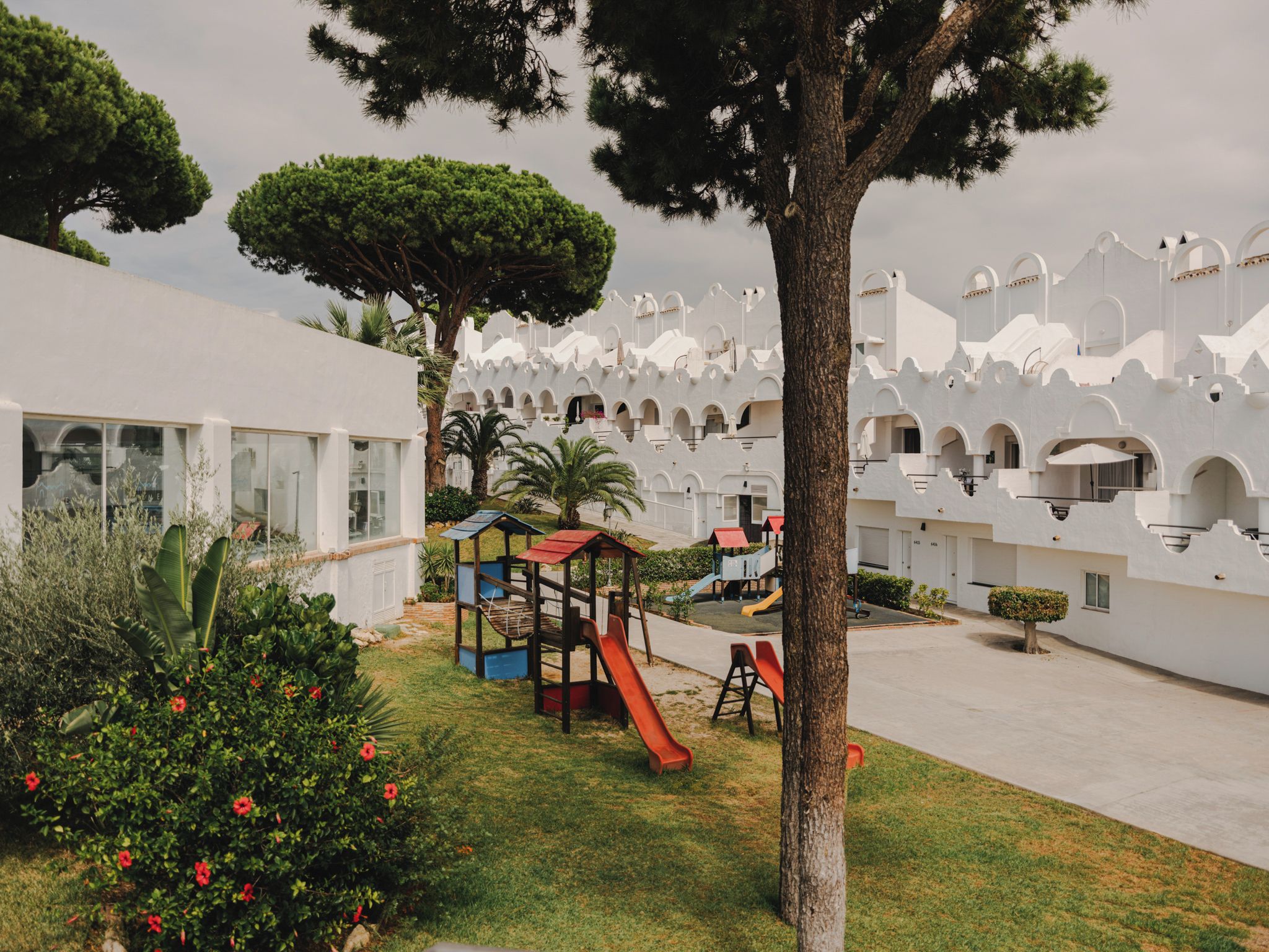 A courtyard with a playground, palm trees, and a white building with arched windows.