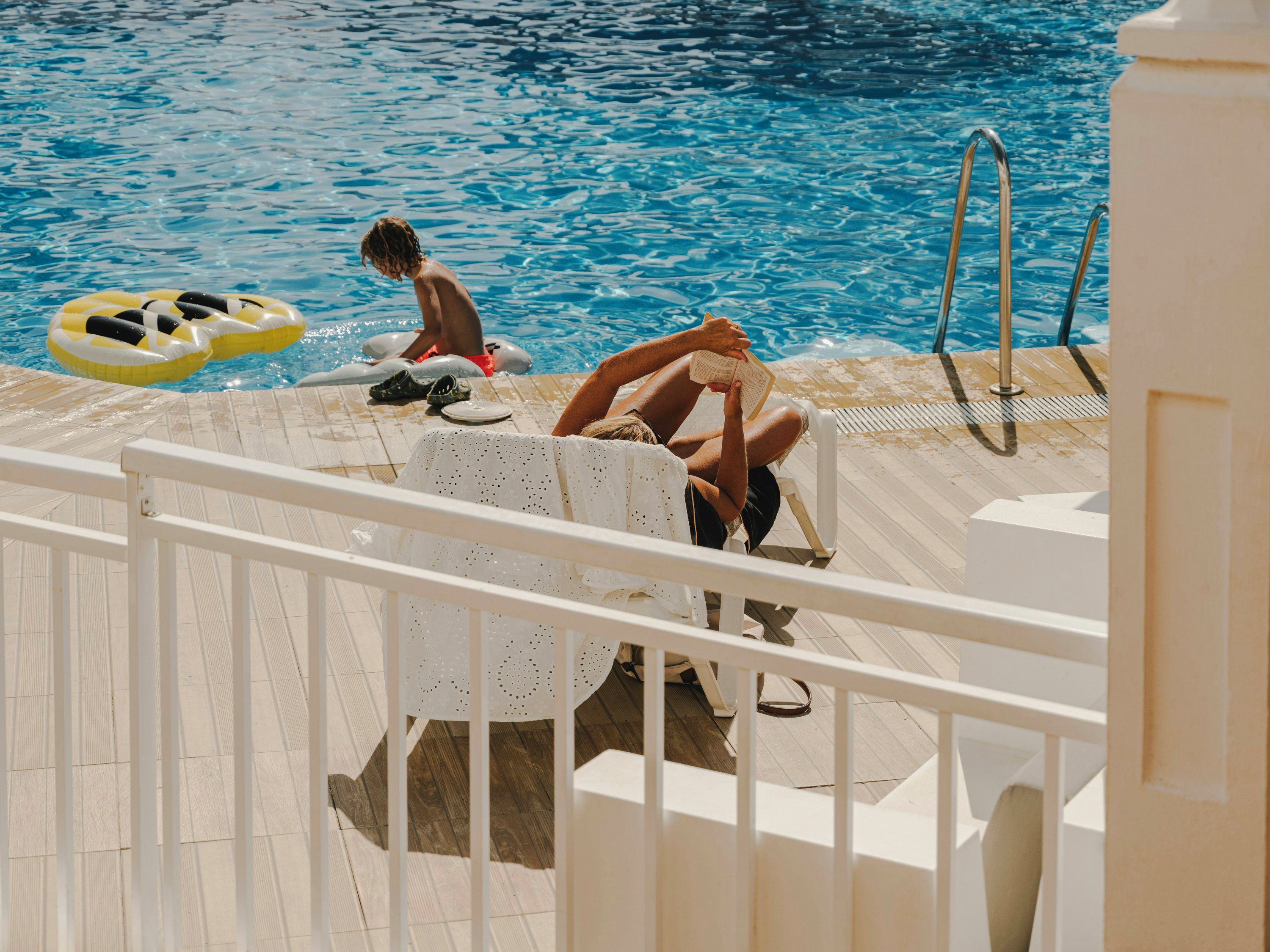 A pool area with a white deck, a pool ladder, and a person reading a book.