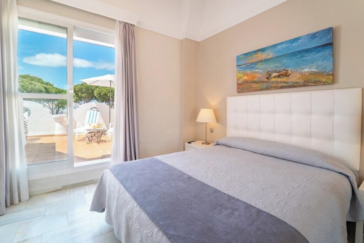 Un cuarto de hotel con una cama, un cuadro, una mesita de noche, una lámpara y vistas al exterior.