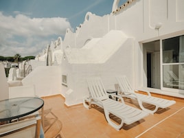 Una terraza con paredes blancas, una mesa de vidrio y dos sillones blancos.