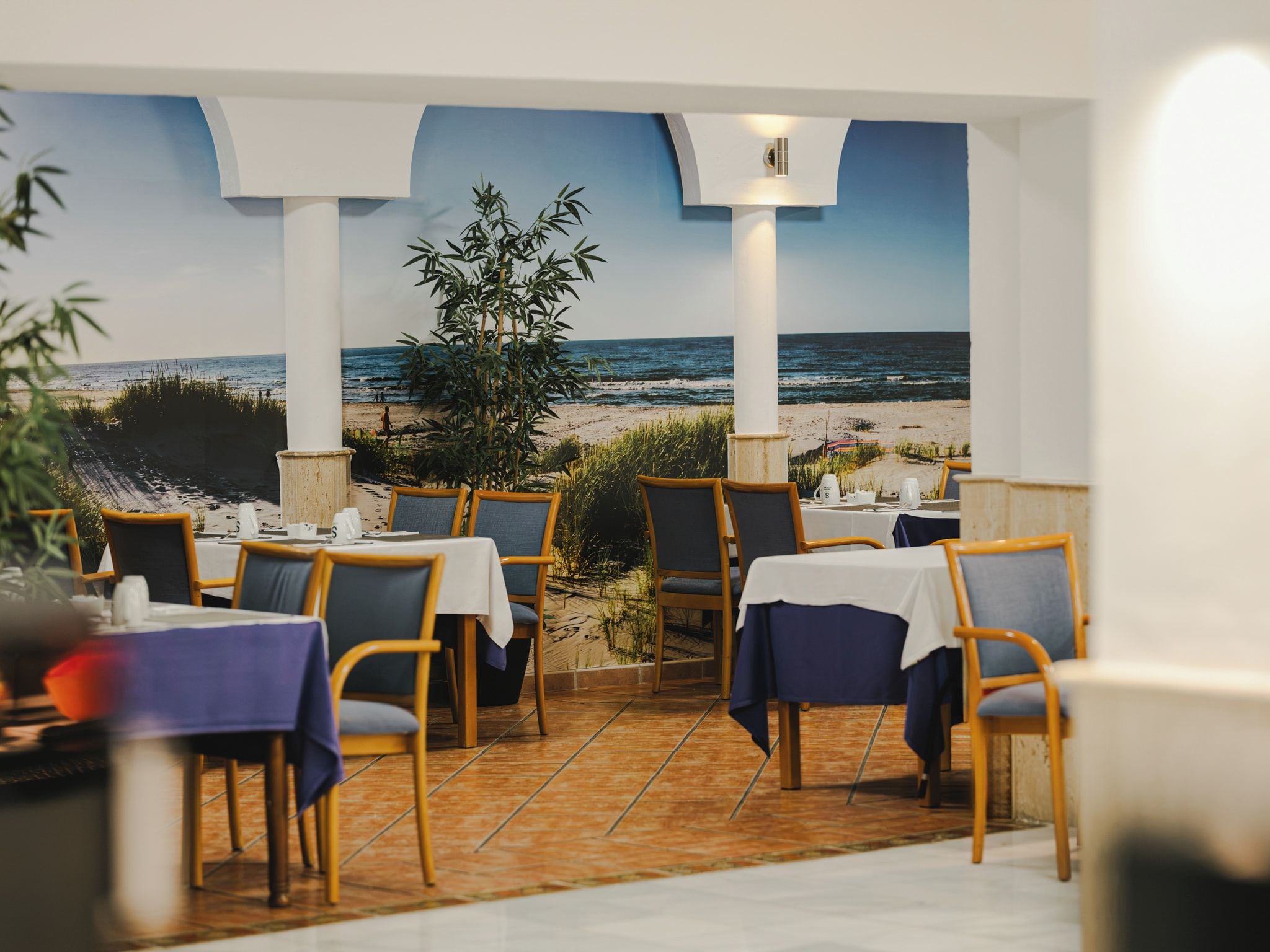 A restaurant with outdoor seating overlooking a beach, featuring tables and chairs arranged on a tiled floor.