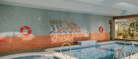 An indoor swimming pool with two separate sections, a tiled wall with a mosaic design, and a window providing a view of greenery.