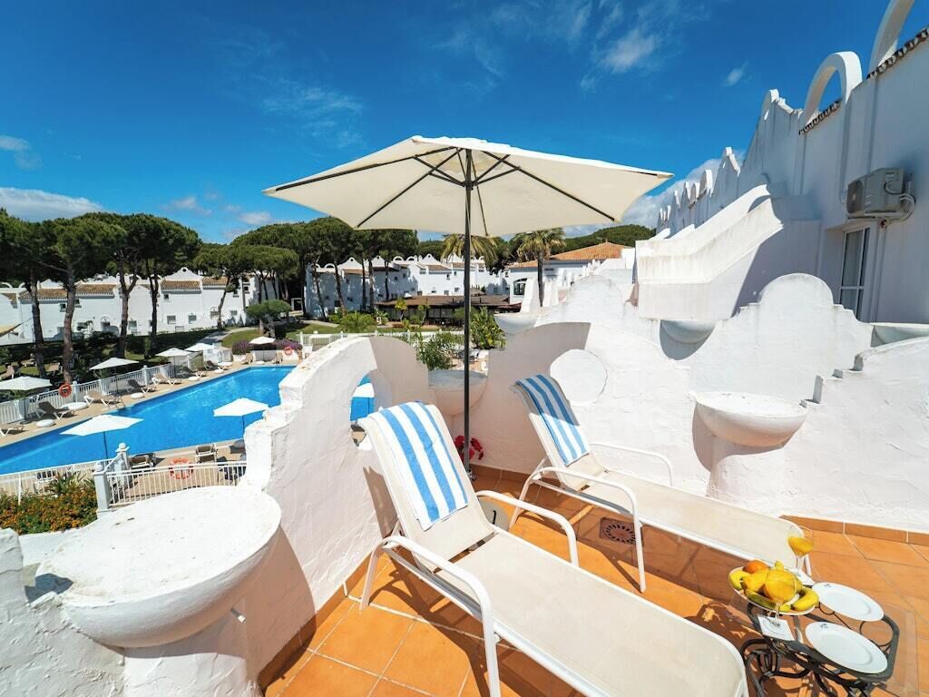 A rooftop pool area with lounge chairs, a table with fruit, and a view of the pool and surrounding buildings.
