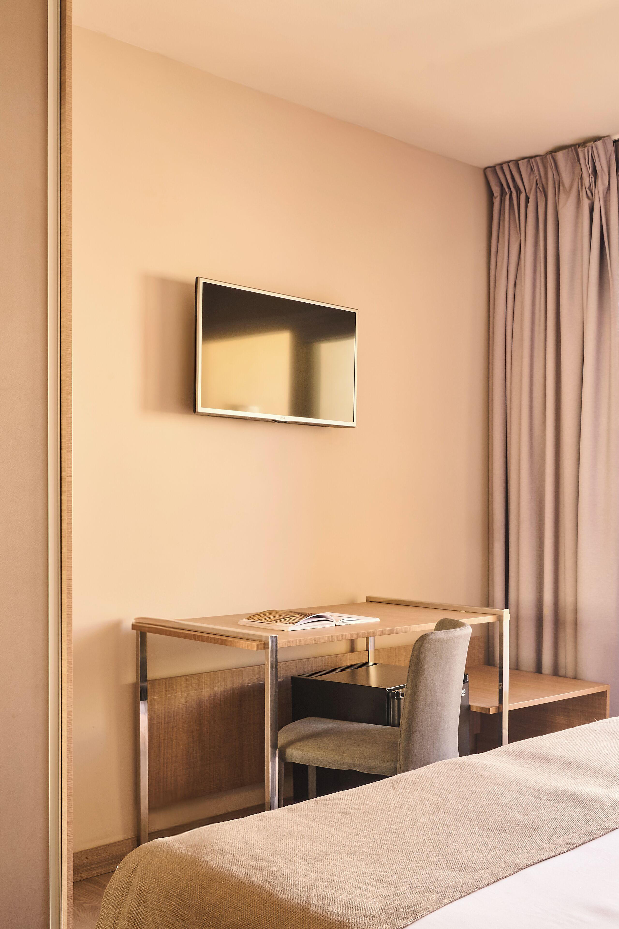 Habitación de hotel con cama, escritorio con silla, televisor en la pared y ventana con cortinas.