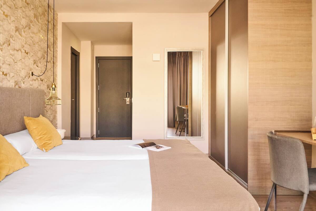 A hotel room with a large bed, a desk, and a chair. There is a door leading to another room and a mirror on the wall.