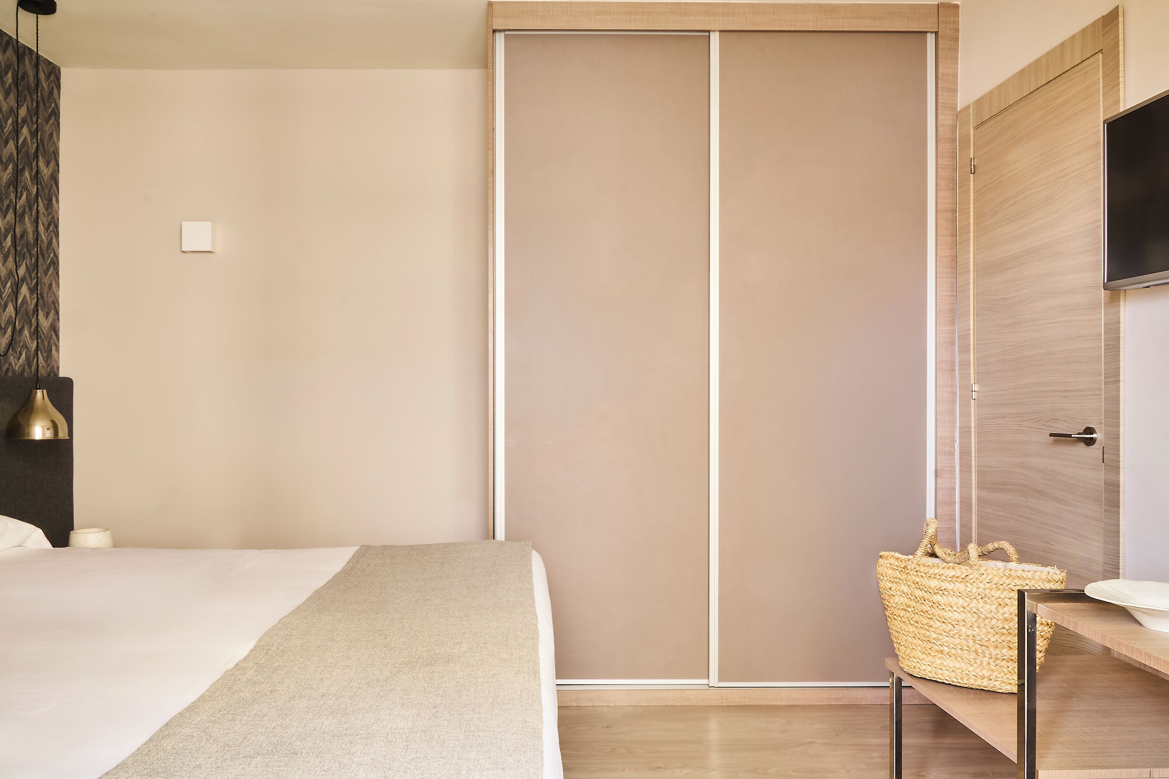 Ein Schlafzimmer mit Bett, Nachttisch, einem gewebten Korb, einem an der Wand montierten Fernseher und einer Schiebetür.