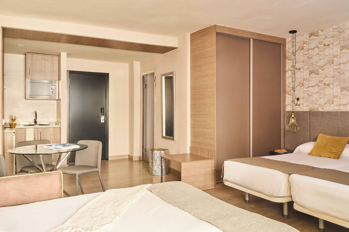 A modern hotel room with a bed, a desk, and a chair. There is a microwave and a small table with a glass top.