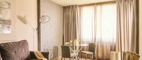 Een moderne woonkamer met een bruine bank, een glazen eettafel met stoelen, een vloerlamp en een groot raam met gordijnen.