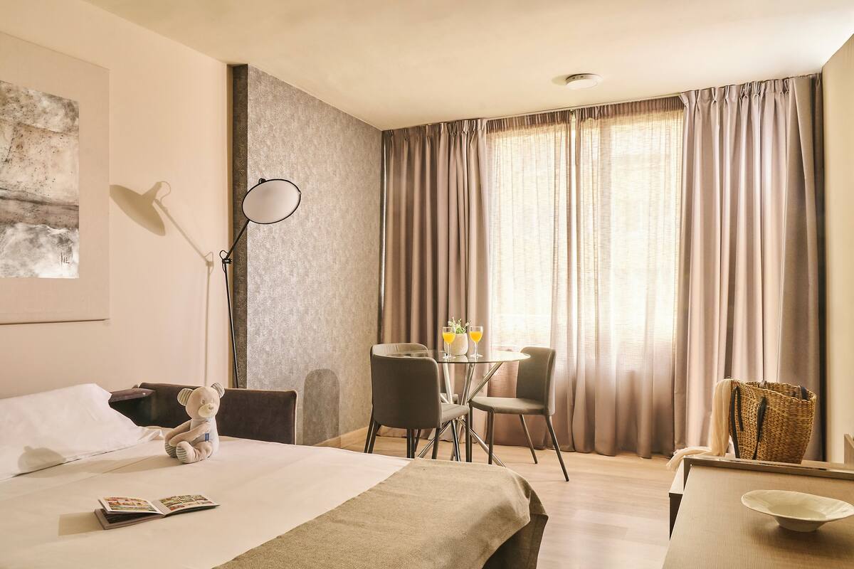 A hotel room with a bed, a desk, a chair, a lamp, a table with two glasses of water, and a teddy bear.