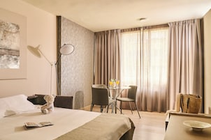 Een hotelkamer met een bed, een bureau, een stoel, een lamp, een tafel met twee glaasjes water, en een teddybeer.