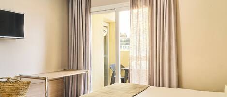 Une chambre d’hôtel équipée d’un lit, d’un bureau, d’une télévision et offrant une vue sur l’extérieur grâce à une fenêtre avec des rideaux.