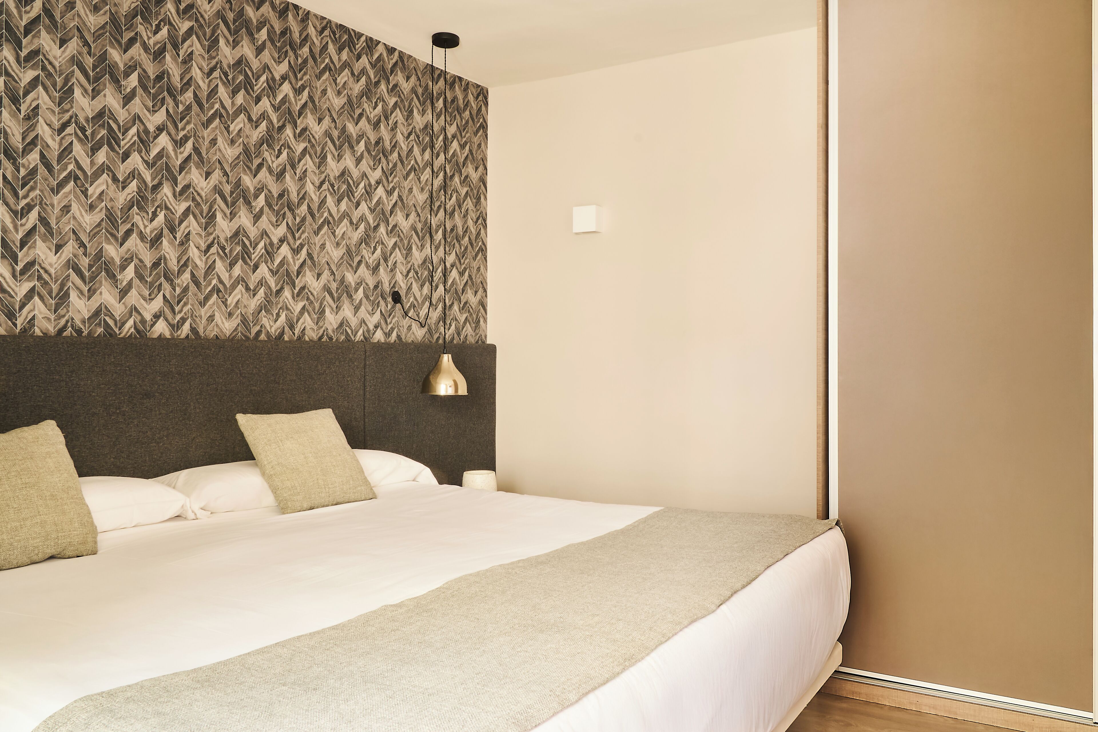 A neatly made bed with a grey headboard, a bedside lamp, and a patterned wallpaper on the wall.
