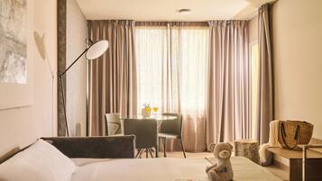 Habitación de hotel con una cama, un escritorio con silla, una lámpara, un osito de peluche y una ventana con cortinas.