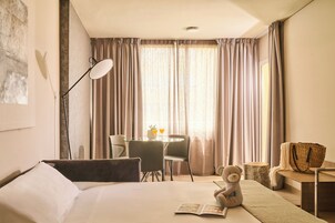 Een hotelkamer met een bed, bureau met stoel, lamp, een teddybeer en raam met gordijnen.