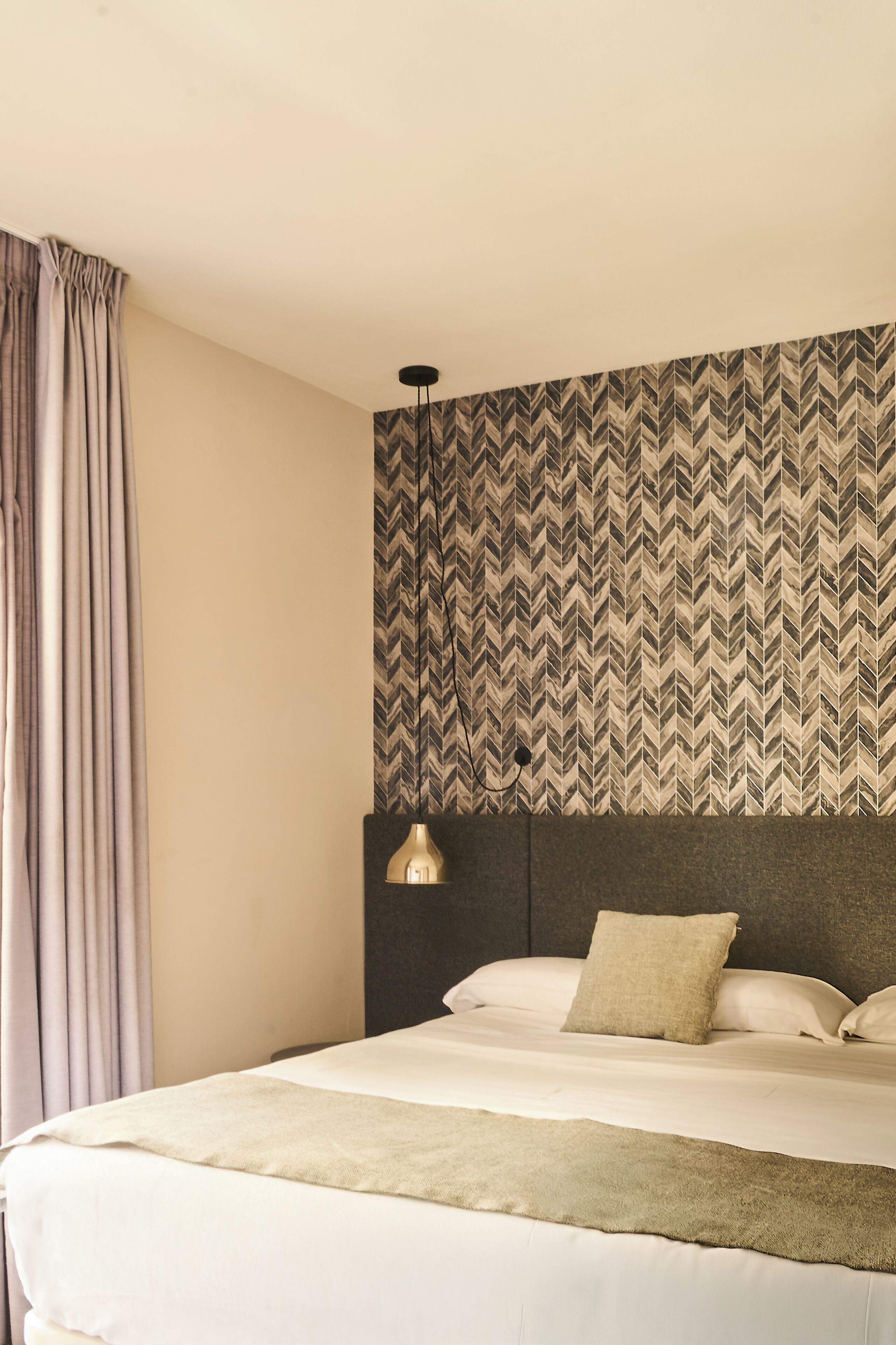 Ein Hotelzimmer mit Bett, Vorhängen, einer Wand mit Muster und einer Pendelleuchte.