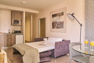 Een moderne hotelkamer met een bed, een kleine kitchenette, een zithoek met tafel en stoelen, en een groot raam.