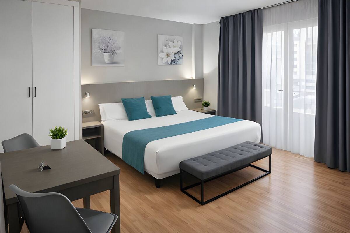 A modern hotel room with a large bed, a bedside table, a desk with a chair, and a window with curtains.